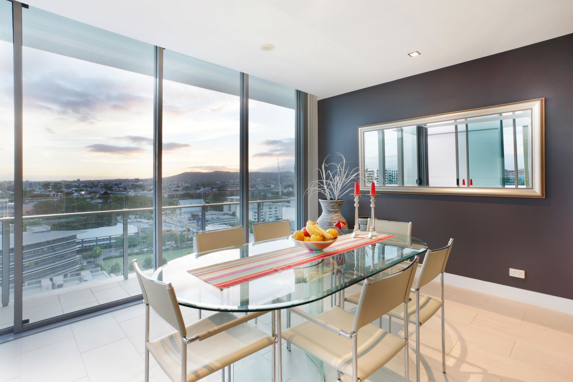 Modern dining room with glass table, white chairs, and panoramic city view through floor-to-ceiling windows