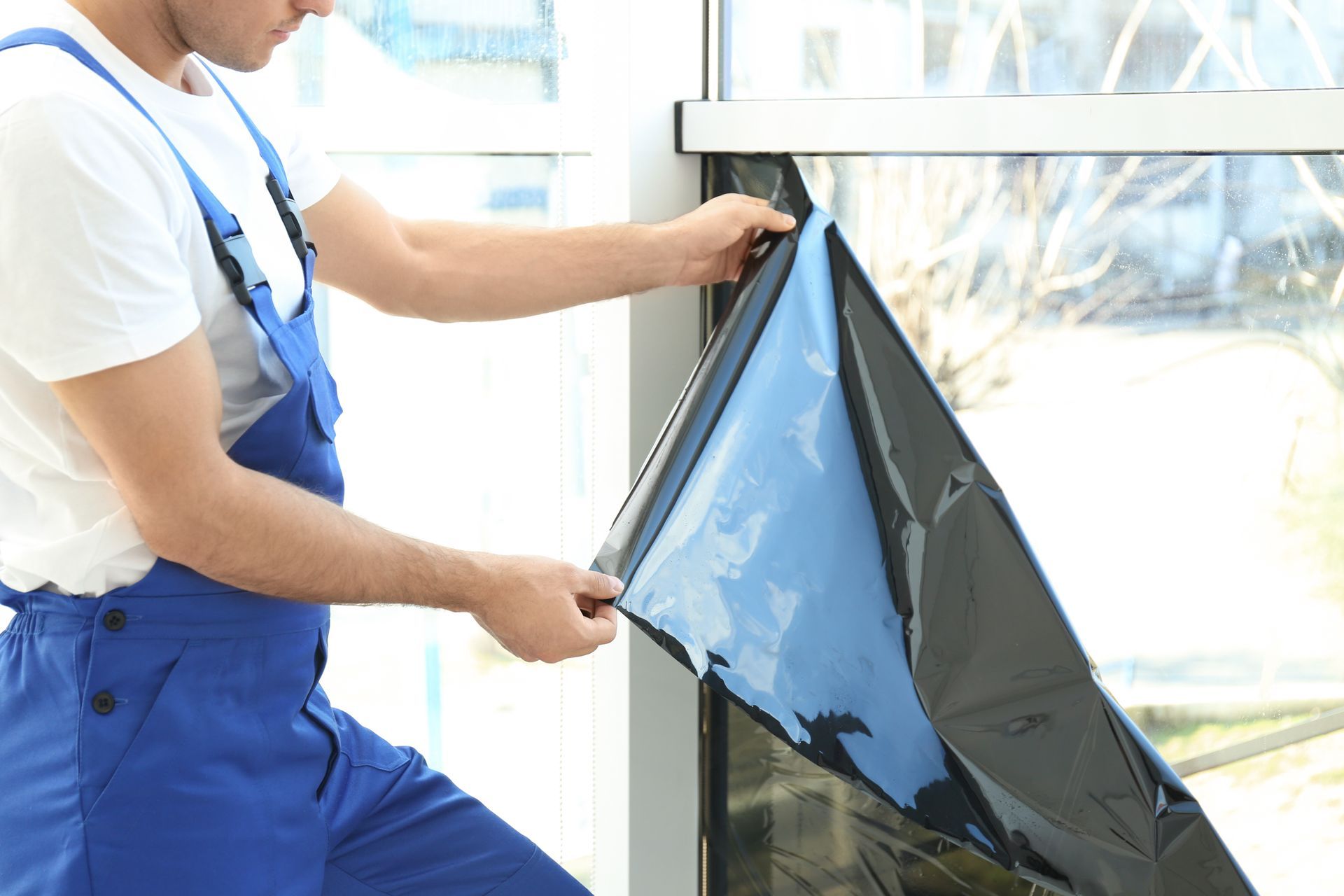 Person in blue overalls fitting a dark window film to a glass pane indoors