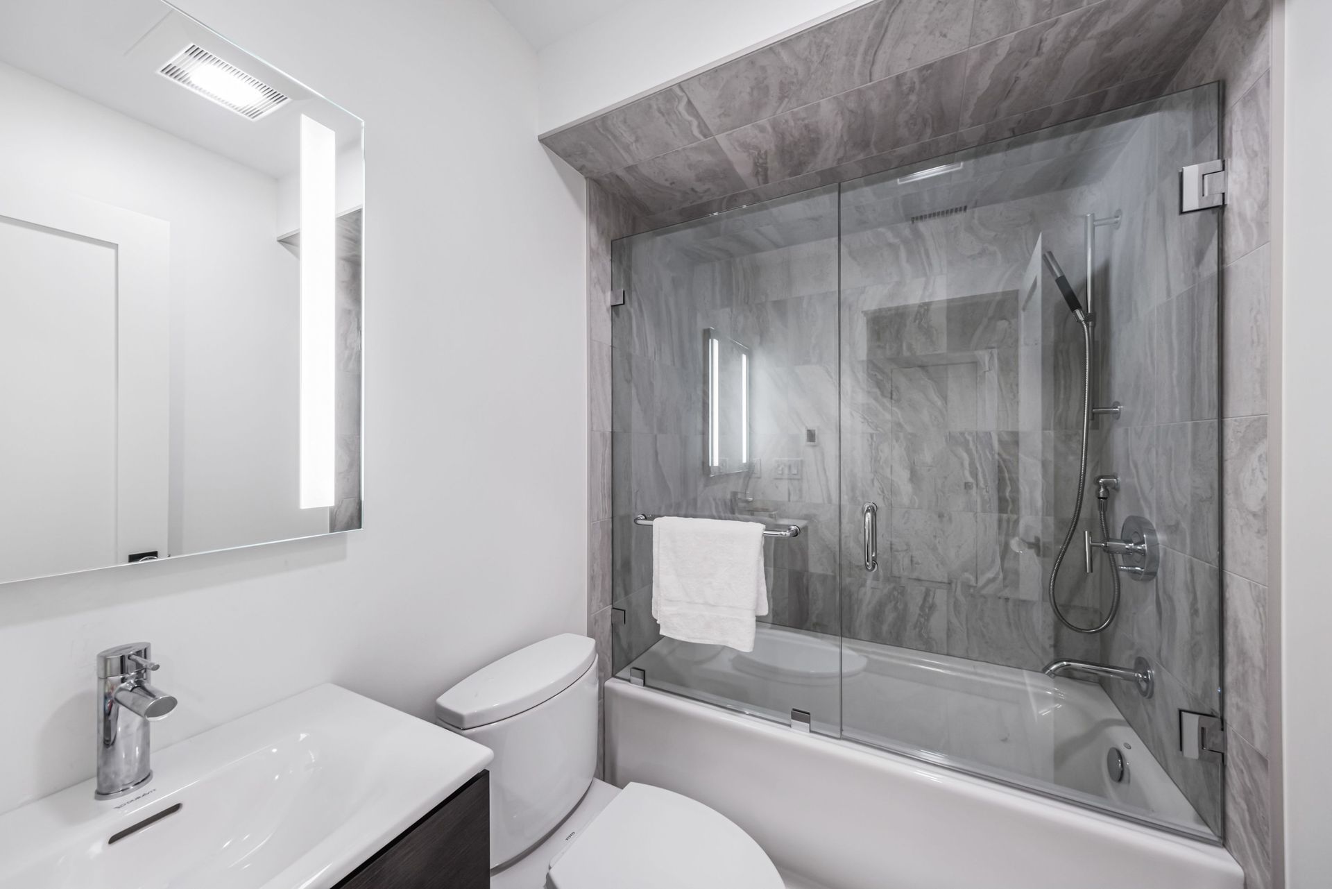 Modern white bathroom with vanity, toilet, and a bathtub enclosed by a glass shower screen.