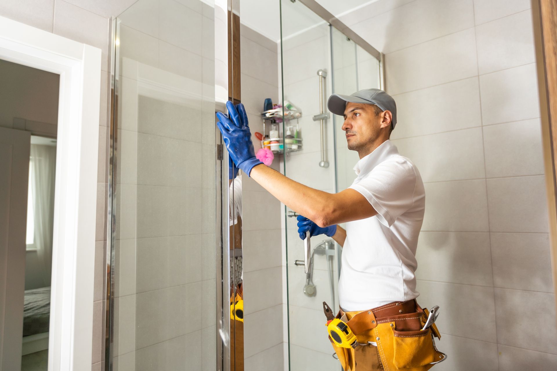 Technician in white shirt and tool belt installing a shower fixture in a tiled bathroom.