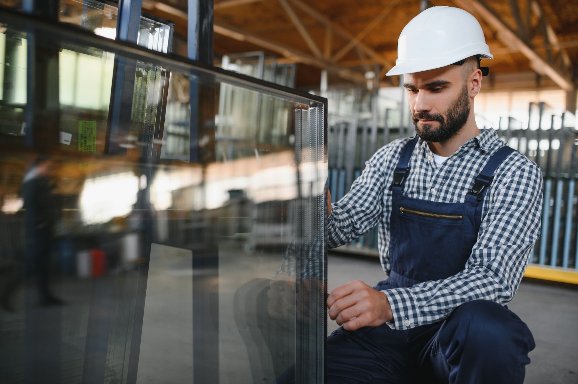 Worker in white hard hat inspecting a large glass pane in a workshop.