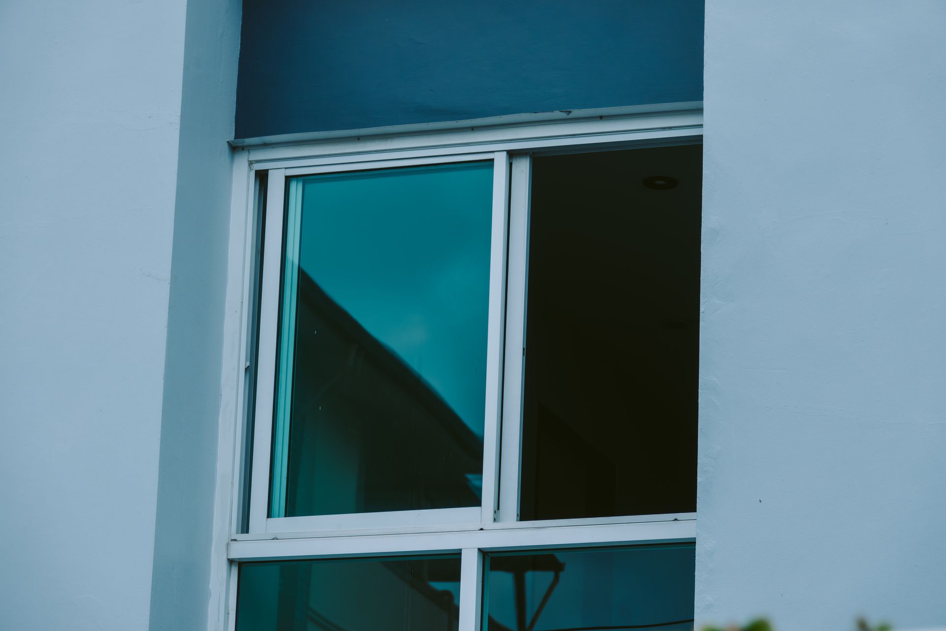 White-framed window in a blue building wall, with dark glass reflecting a slanted roof.