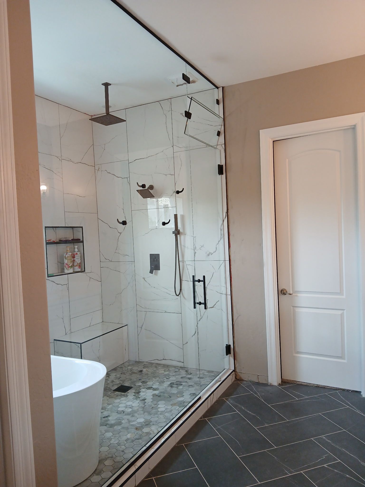 Modern bathroom with glass shower, white tub, and dark hexagon tile floor.