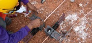 Workers installing electrical grounding components on a dirt surface, with copper wires and safety gear.