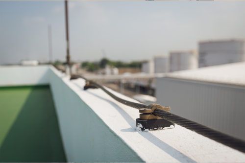 A black cable secured by clamps runs along a white rooftop wall, with blurred industrial buildings in the background.