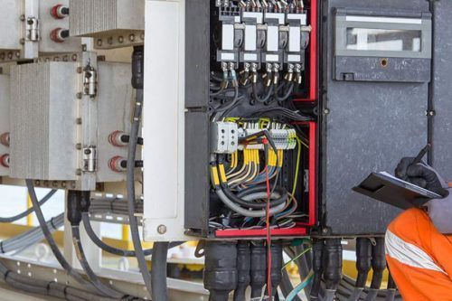 A person in orange overalls inspecting electrical panels with wires and gauges.