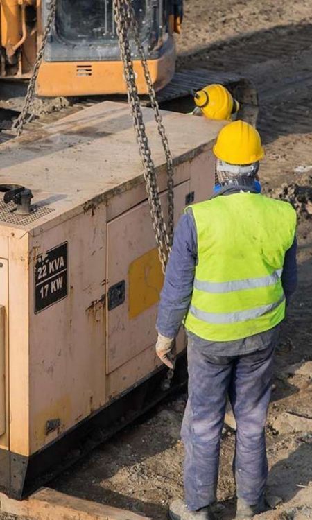 Construction worker in yellow vest and hard hat watches a generator being lifted by a machine at a construction site.