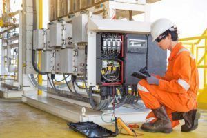 Electrician in orange jumpsuit inspects electrical equipment, outdoors.