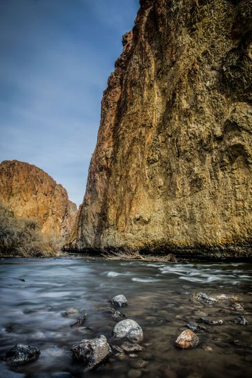 River flows past textured rock formations under a blue sky.