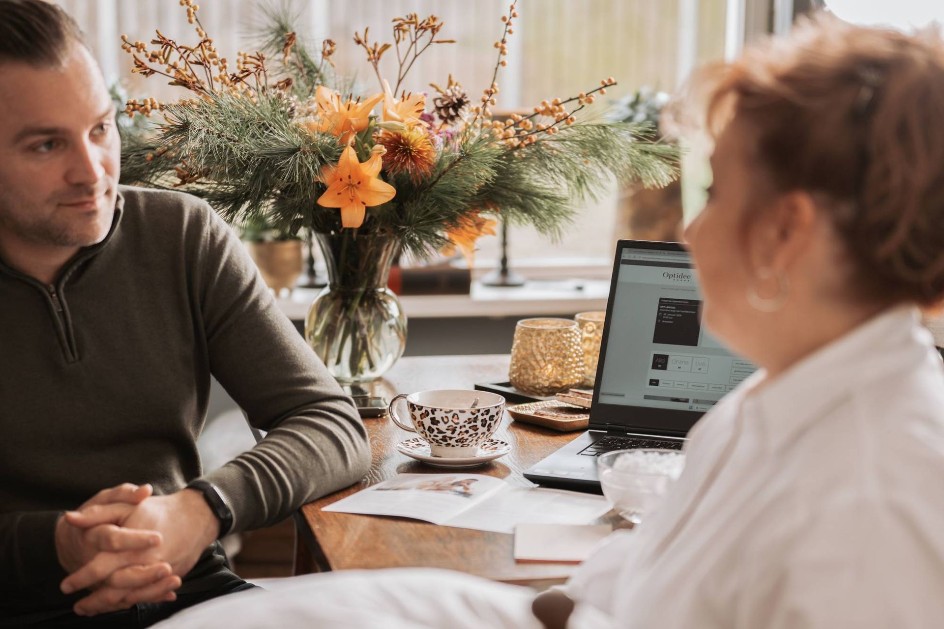 Een man en een vrouw zitten aan een tafel met een laptop, bloemen en een kop koffie; de man kijkt naar de vrouw.