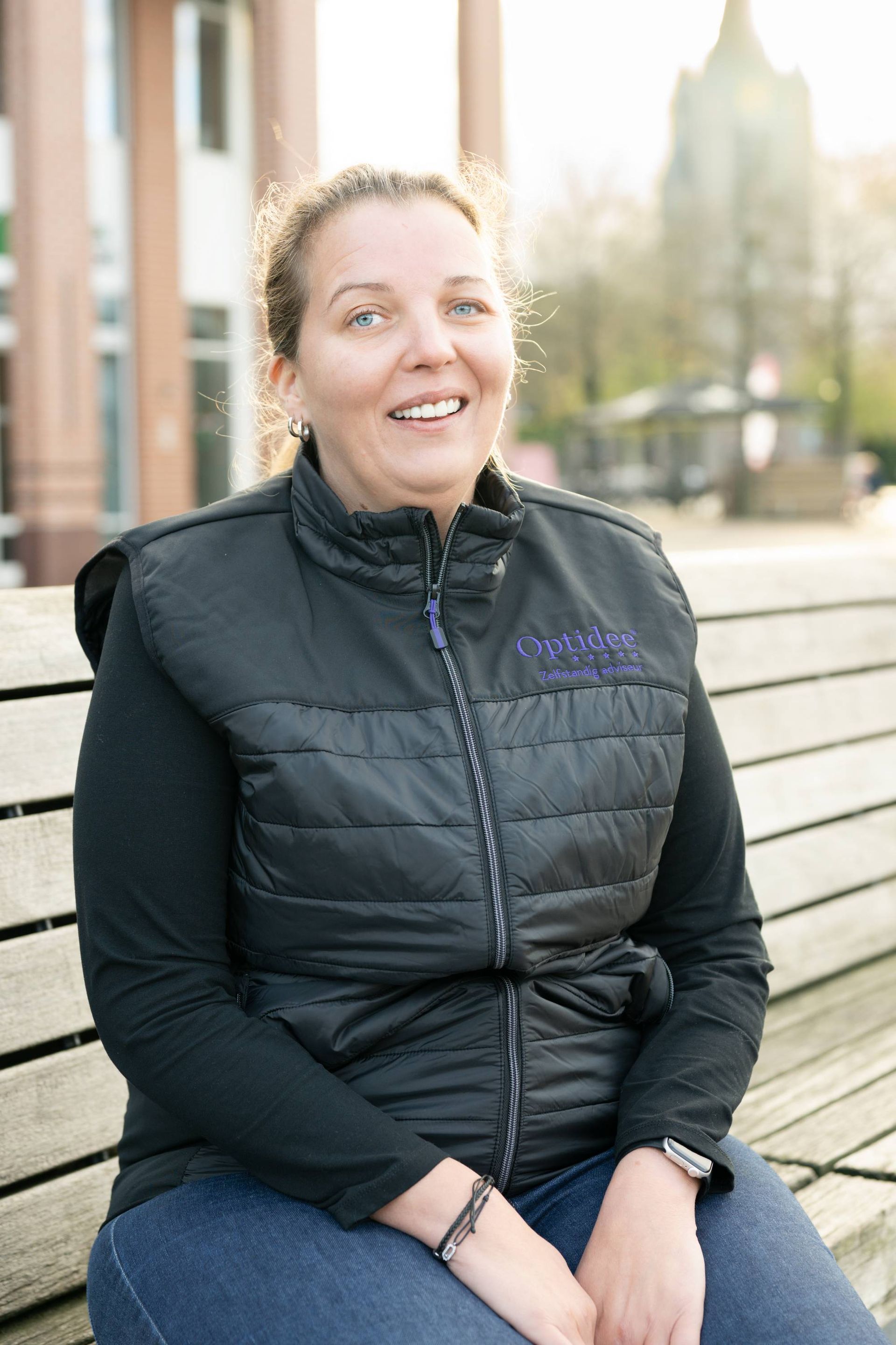 Vrouw in zwart vest glimlacht, zittend op een bankje buiten. Gebouw op de achtergrond.