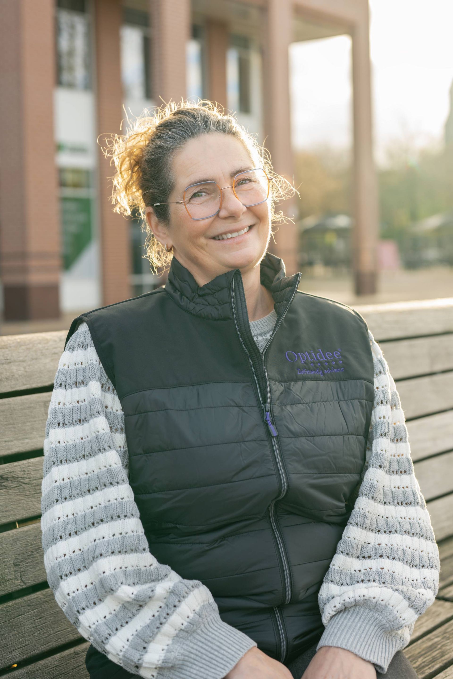 Een vrouw in een vest en met een bril zit glimlachend op een houten bankje buiten.