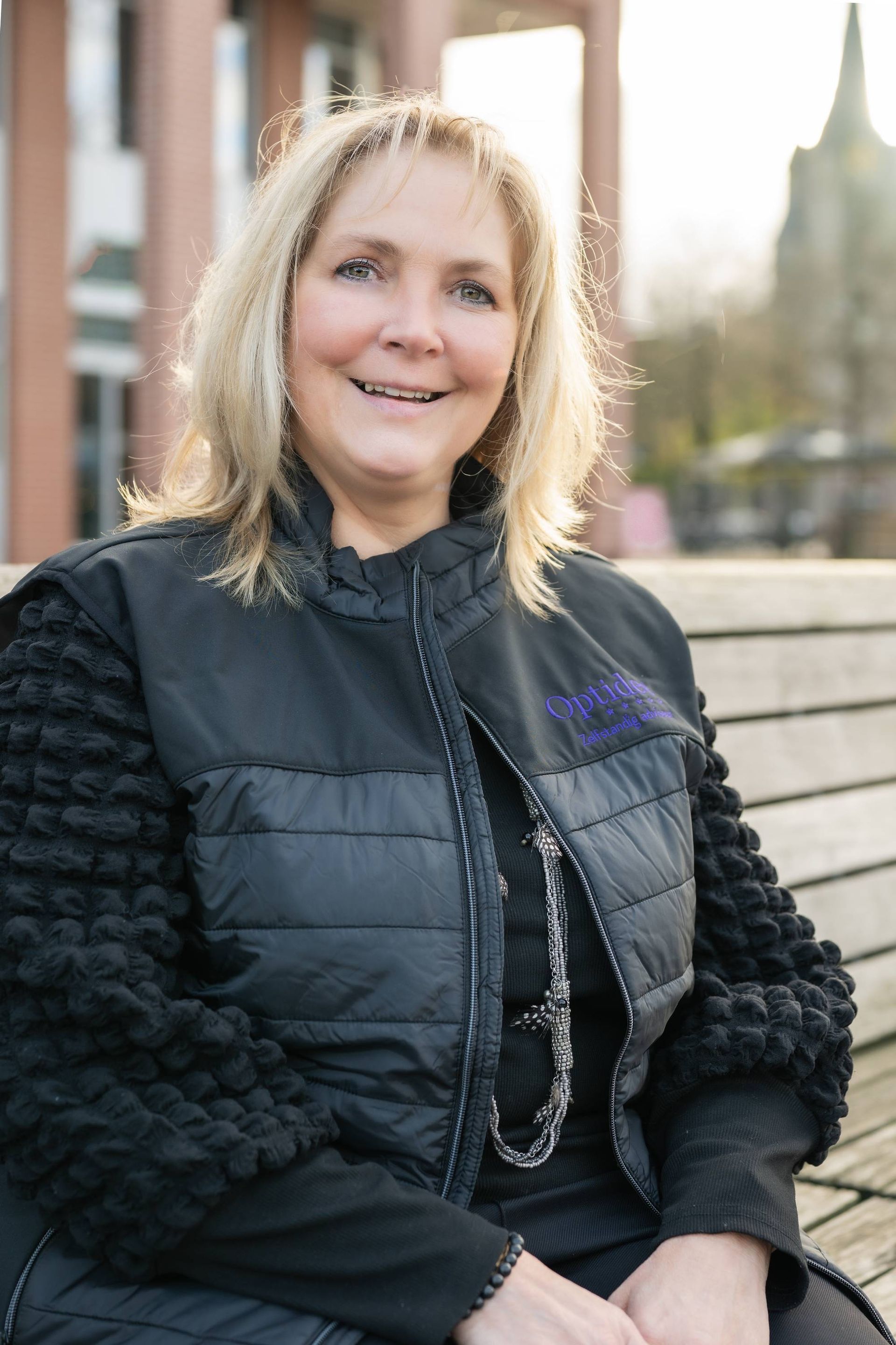 Vrouw met blond haar, gekleed in een zwart vest, zit lachend buiten met een gebouw op de achtergrond.