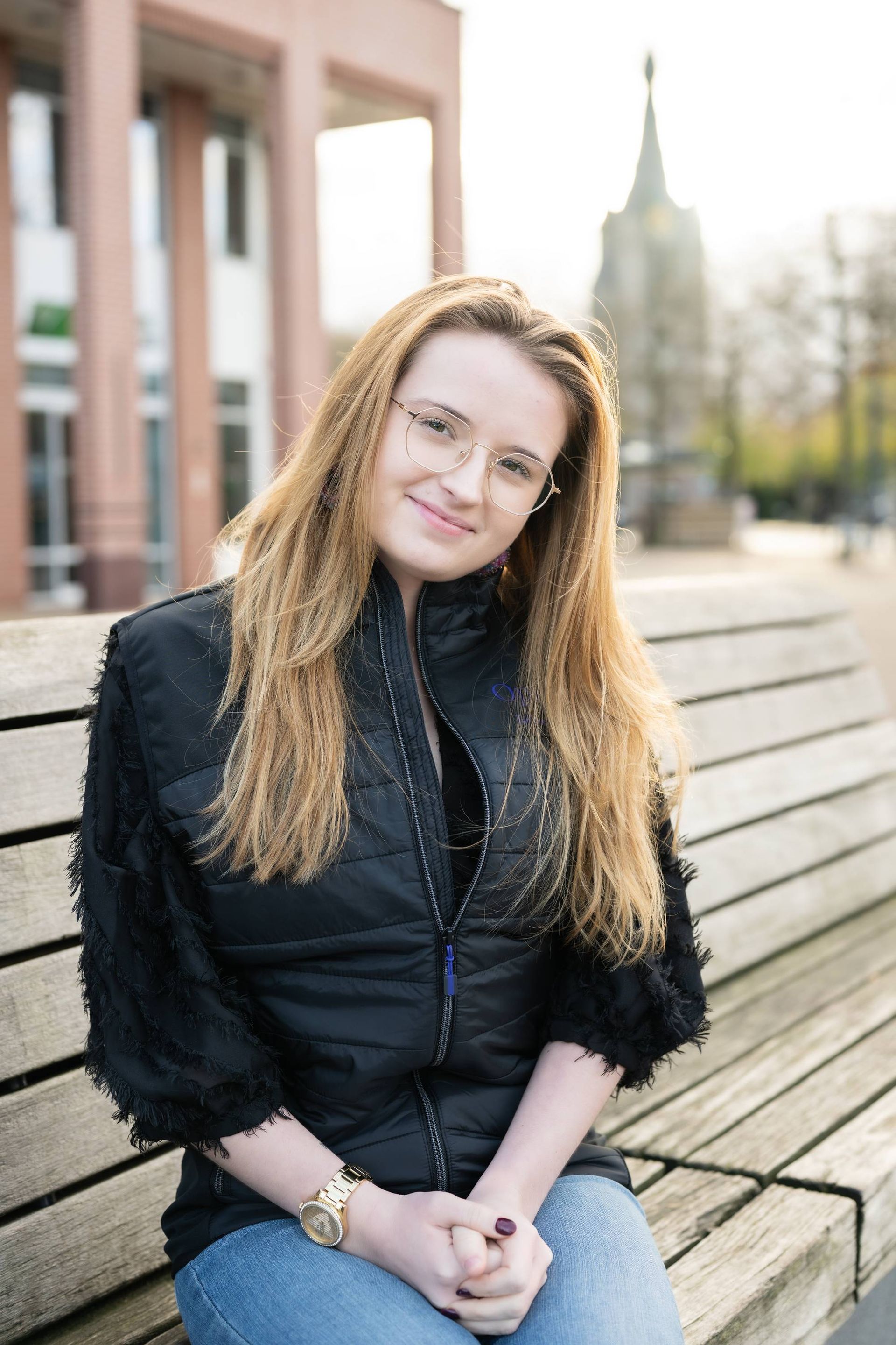 Een vrouw met een bril zit op een parkbankje, gekleed in een zwarte jas en spijkerbroek.