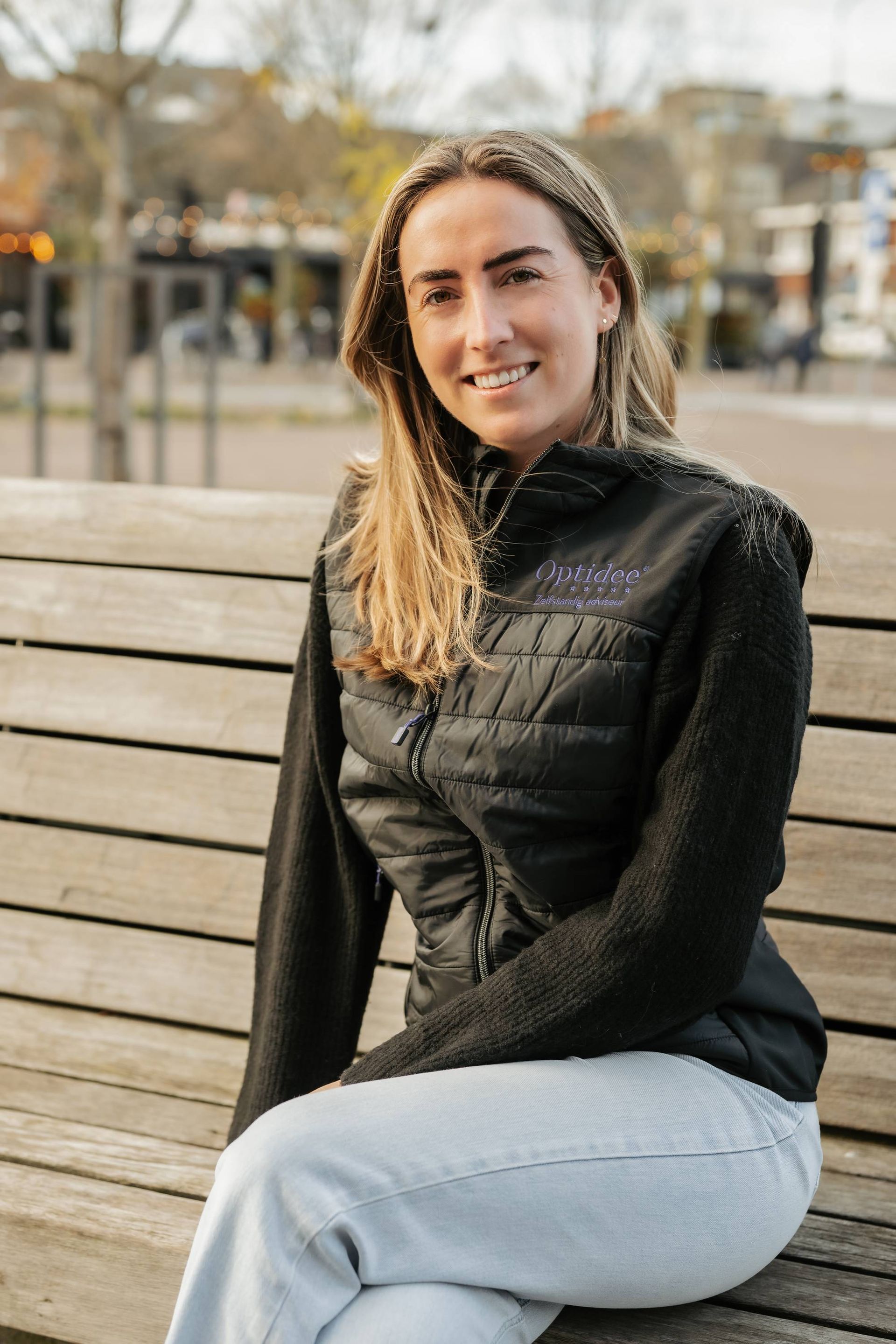 Vrouw in zwart vest en lichte spijkerbroek, lachend, zittend op een houten bankje buiten.