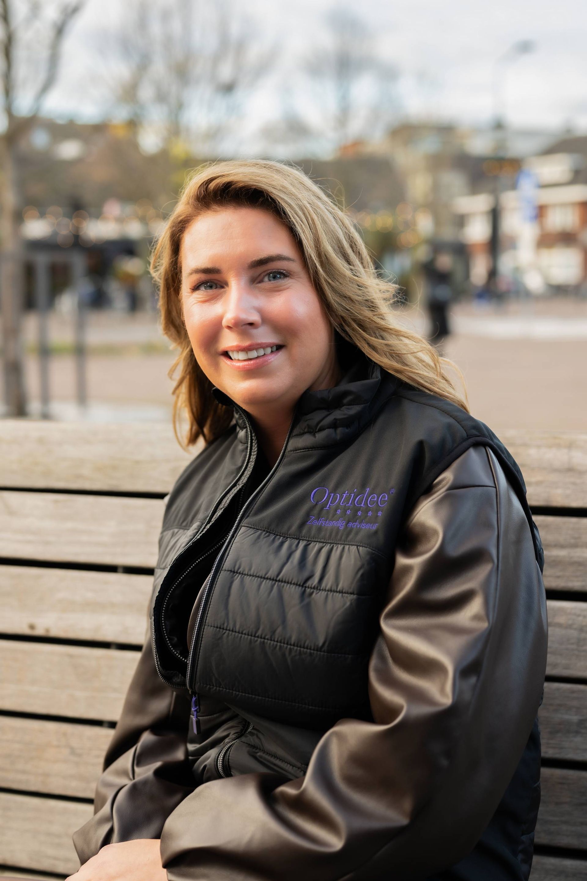 Een vrouw in een zwart gewatteerd vest met leren mouwen zit lachend op een houten bankje in de buitenlucht.