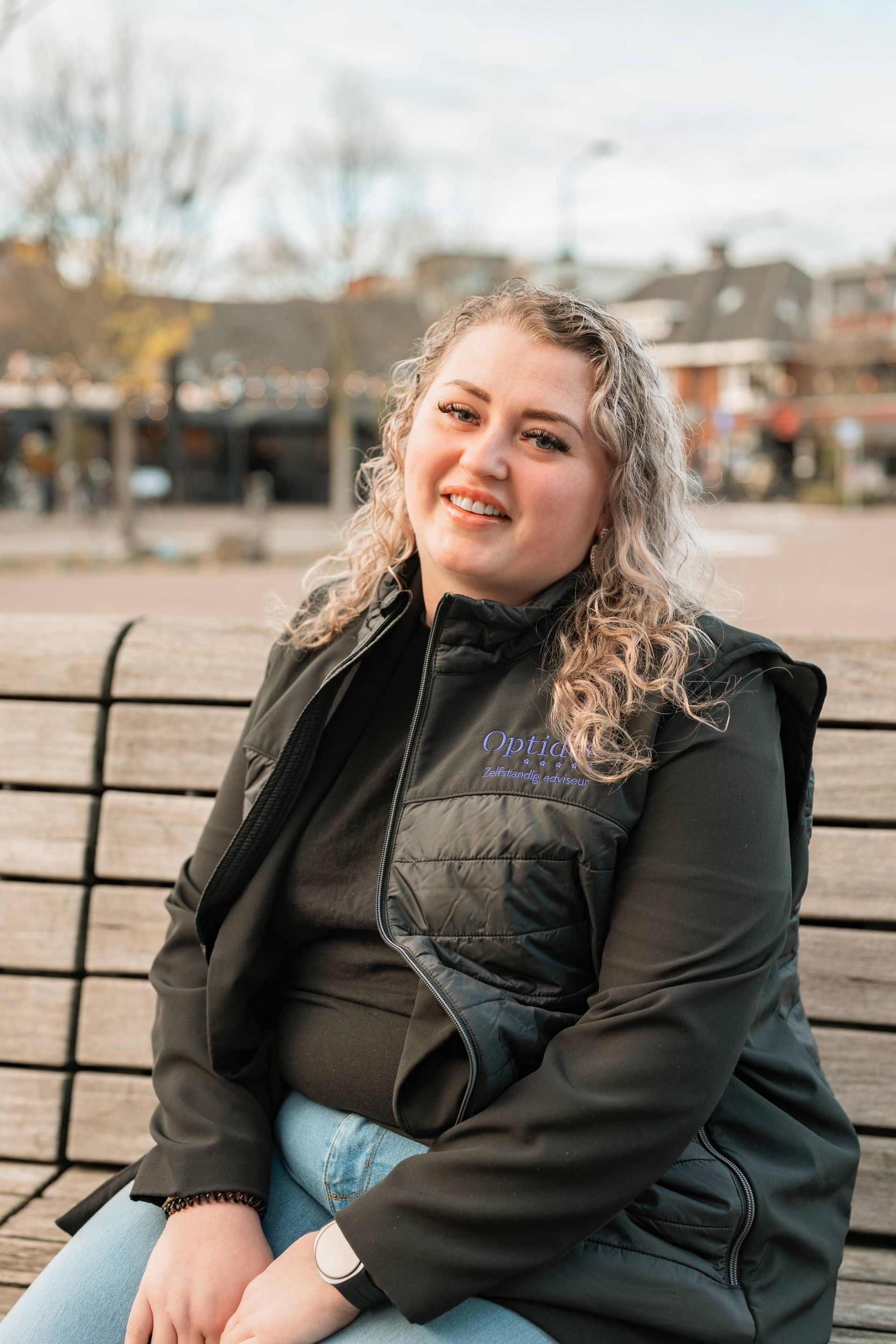 Een vrouw met krullend blond haar glimlacht, zittend op een parkbankje, gekleed in een zwart vest en lichtblauwe broek.