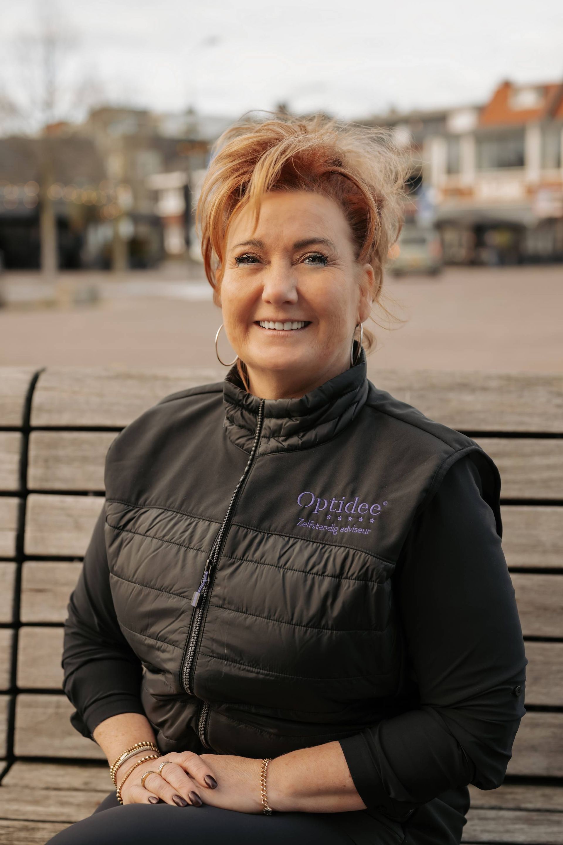 Vrouw met roodblond haar in een zwart vest, zittend in de buitenlucht. Lachend.