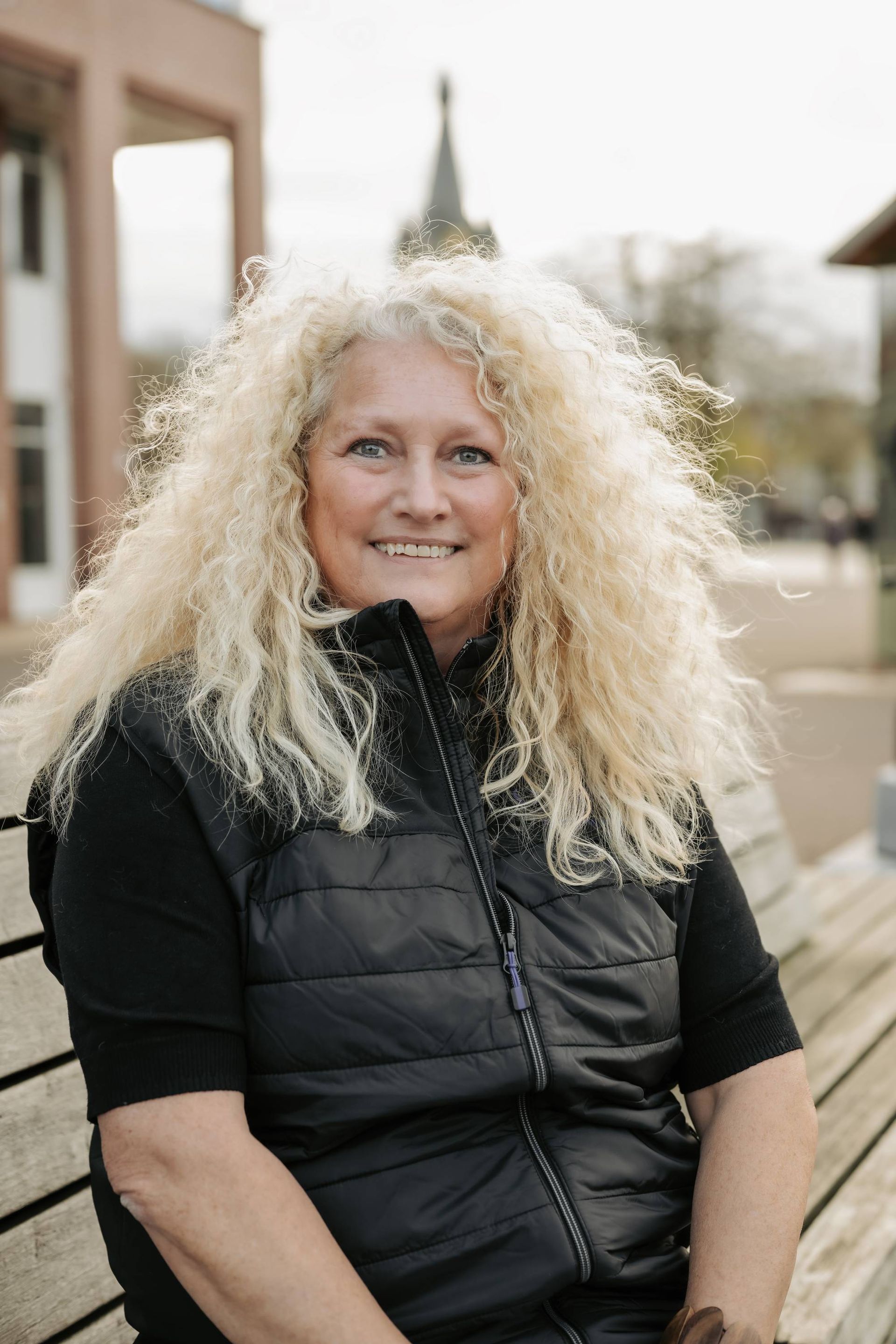 Vrouw met krullend blond haar, gekleed in een zwart vest, zit lachend op een bankje buiten.