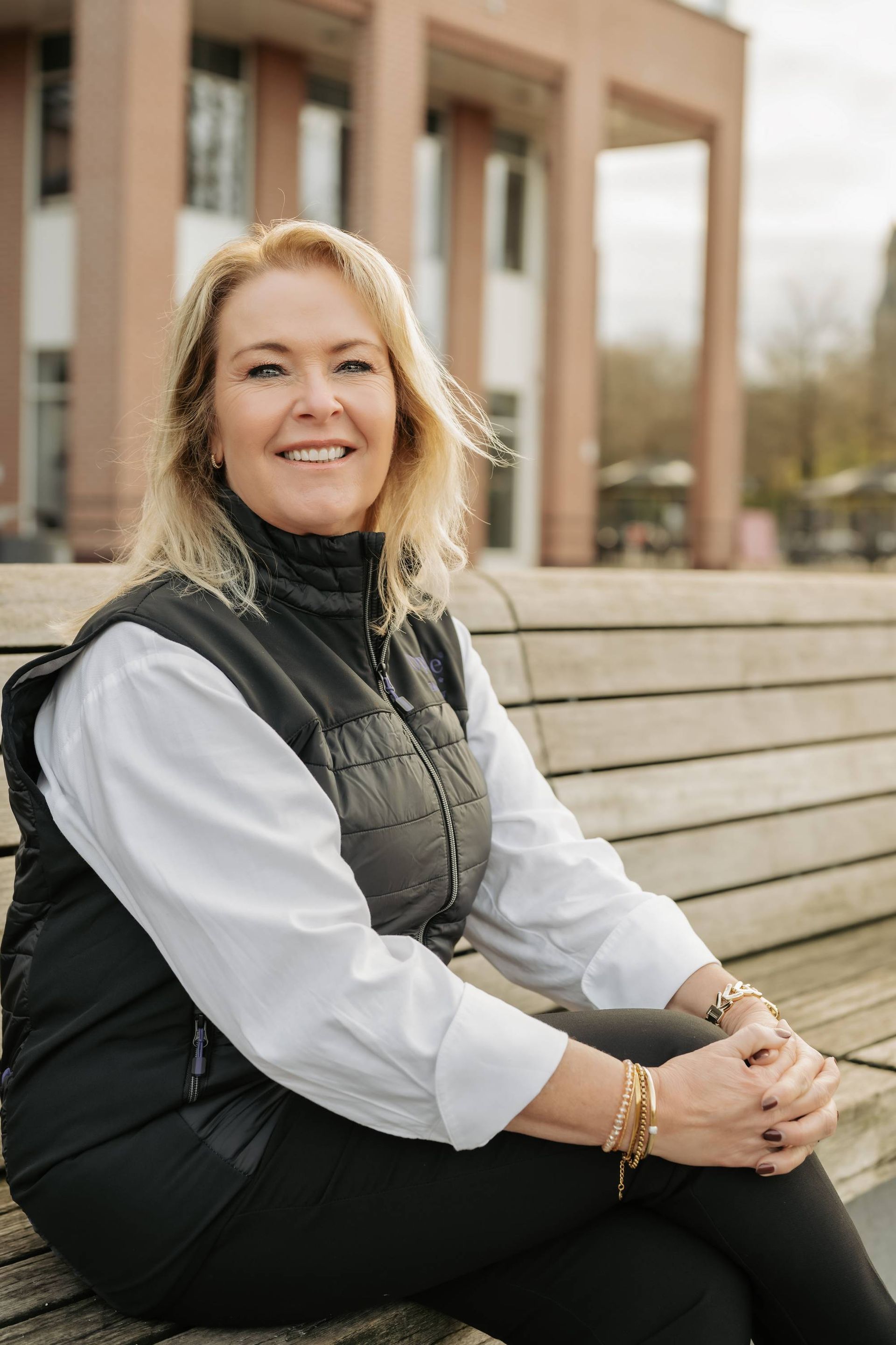 Een vrouw met blond haar, gekleed in een zwart vest en een witte blouse, glimlacht terwijl ze op een houten bankje zit.