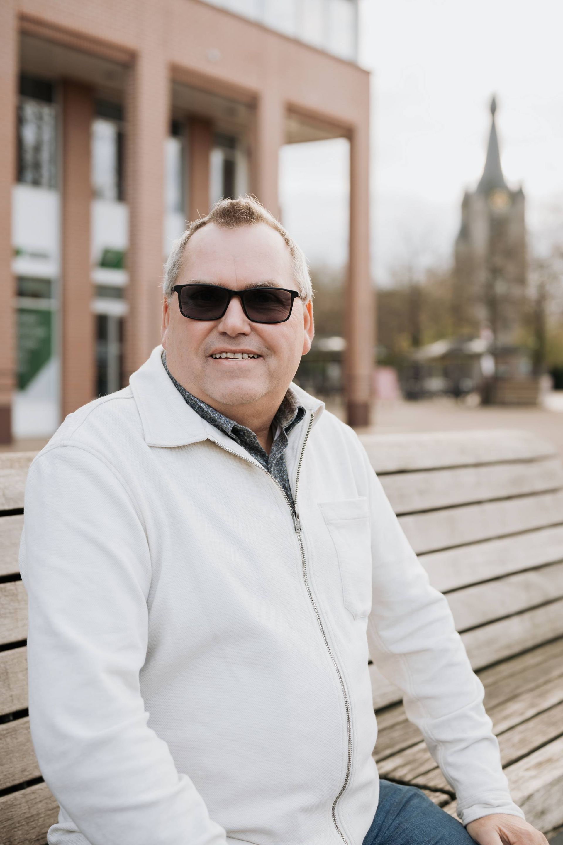 Een man met een zonnebril en een wit shirt zit glimlachend op een bankje buiten. 