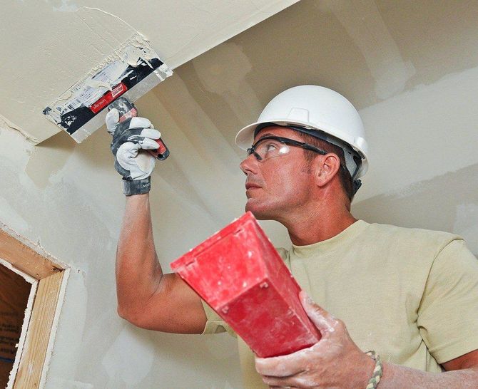 Worker in a hard hat applying plaster to a ceiling with a trowel, holding a red hawk board.
