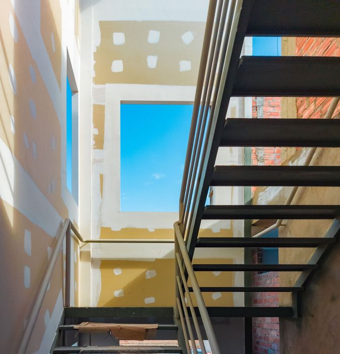 Narrow stairwell with metal stairs and blue sky framed by sunlit cream walls and windows