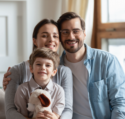 Family of three smiling at the camera, posing indoors; a child holds a stuffed animal.