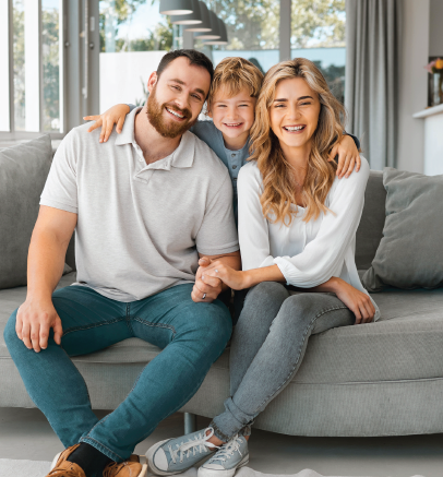 Family of three smiling on a gray couch, arms around each other in a bright living room.
