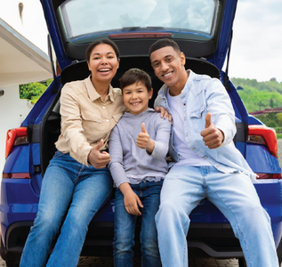 Family of three smiling, giving thumbs-up, sitting in open car trunk. Blue car, sunny day.