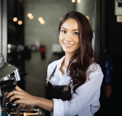 Woman in white shirt and apron operates a coffee machine, smiling in a cafe setting.