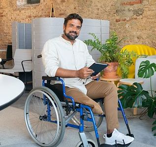 Man in wheelchair holding tablet smiles in an office with plants and exposed brick wall.