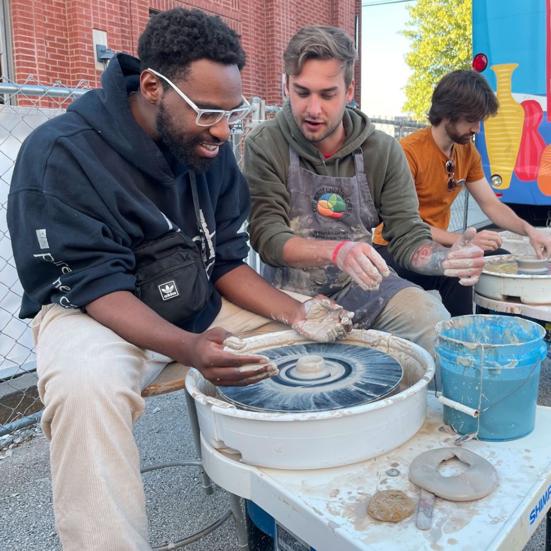 Three people are shaping clay on pottery wheels outdoors, with buckets and clay tools nearby.
