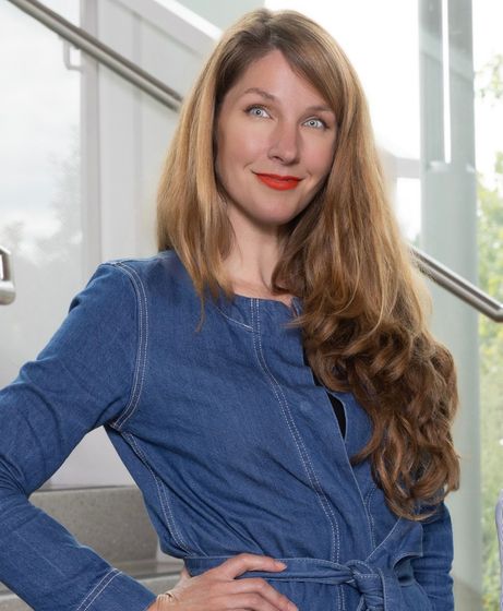 Woman in a blue top posing indoors by a staircase, smiling at the camera.