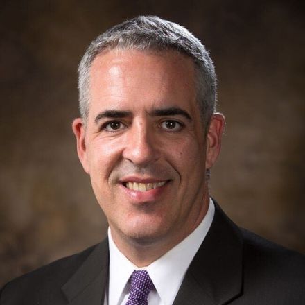 Studio portrait of a smiling man in a dark suit and purple tie on a neutral brown background