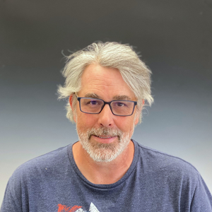 Portrait of a smiling man with gray hair, glasses, and a blue shirt against a gray background