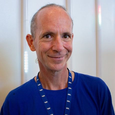 Smiling man in a blue shirt with a lanyard, standing indoors against a light background