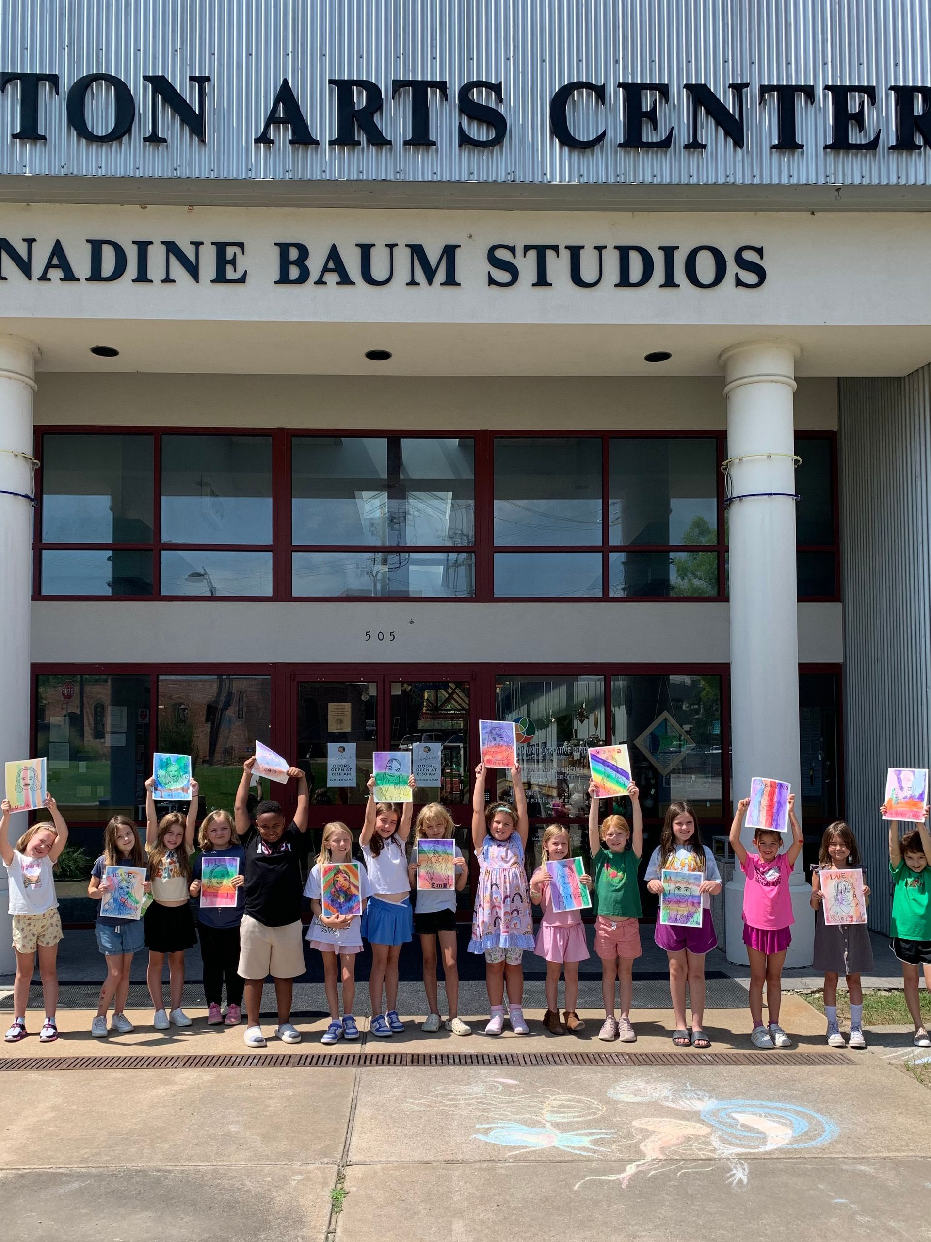 Children holding signs outside the Community Creative Center entrance, with “Nadine Baum Studios” above the doors