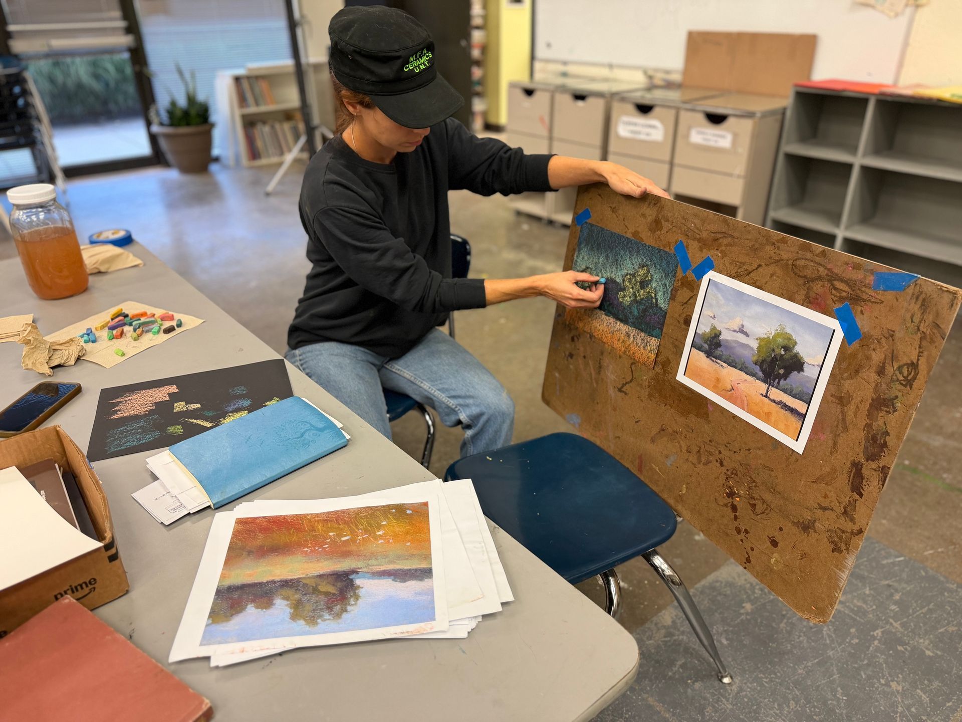 Person arranging artwork on a corkboard beside a table covered with papers and art supplies.