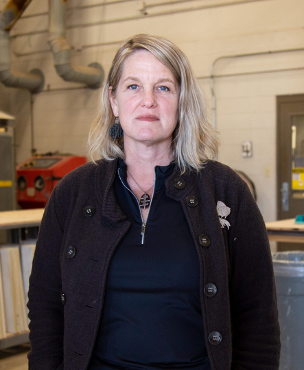 Woman standing in an industrial workshop, wearing a dark jacket and black top, looking at the camera.