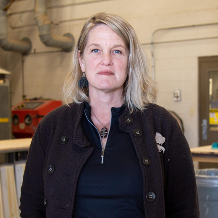 Woman standing in an industrial workshop, wearing a dark jacket and black top, looking at the camera.
