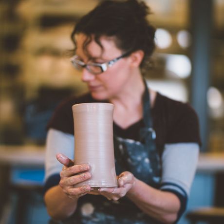 Person holding a tall beige ceramic vase in a workshop, looking down at it