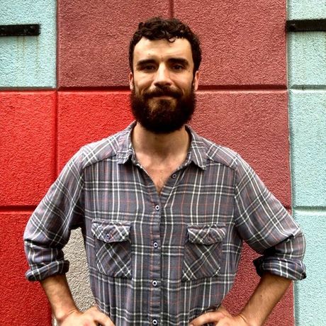 Man with a beard in a plaid shirt standing against a red and gray tiled wall