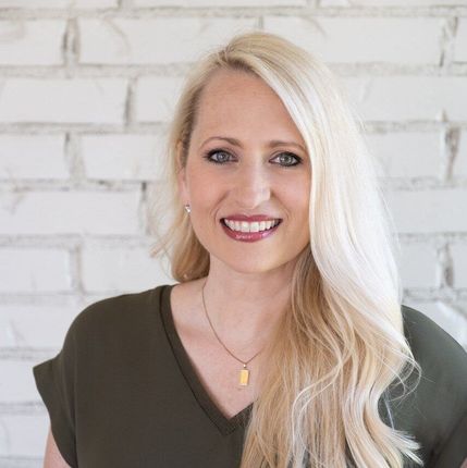 Smiling blonde woman in a dark green top against a white brick wall