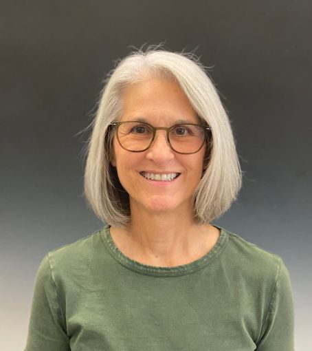 Smiling person with shoulder-length gray hair and glasses, wearing a green shirt against a dark gray background