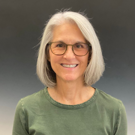 Smiling person with shoulder-length gray hair and glasses, wearing a green shirt against a dark gray background