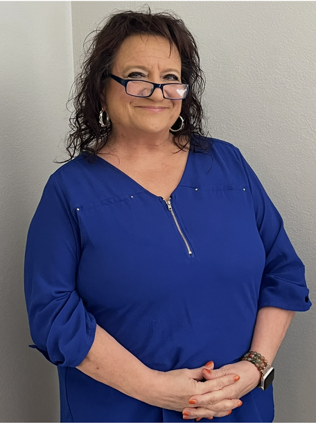 Woman in a blue blouse standing indoors against a plain wall, hands clasped and smiling slightly.