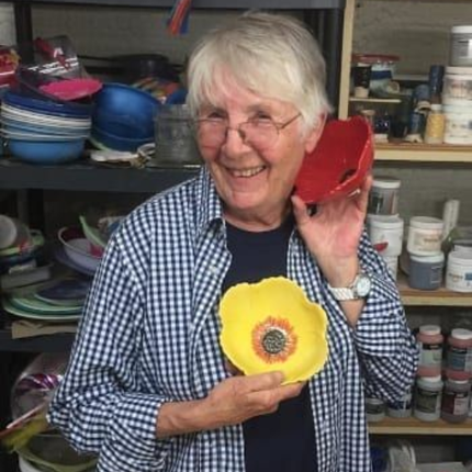 Smiling woman in a shop holding a yellow flower-shaped bowl, with shelves of pottery behind her