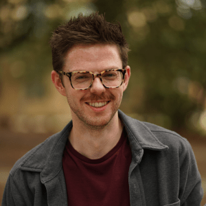 Smiling person with glasses and dark jacket standing outdoors against a blurred background