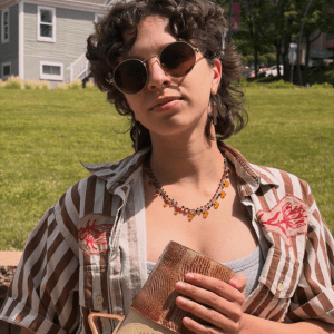 Person wearing round sunglasses and a striped shirt holds a woven clutch outdoors on a sunny lawn